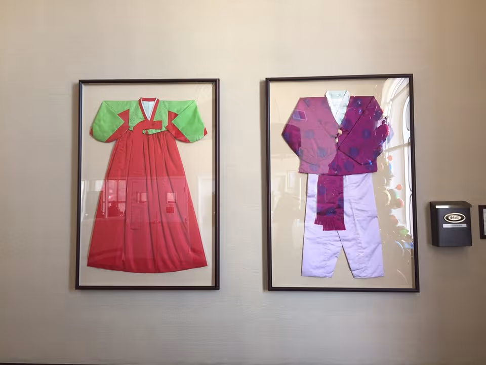 Two framed traditional Korean outfits displayed on a beige wall. The left frame contains a red and green hanbok dress, and the right frame contains a purple jacket with white pants and a purple sash. To the right of the frames, there is a black wall-mounted mailbox labeled 'Mail'.
