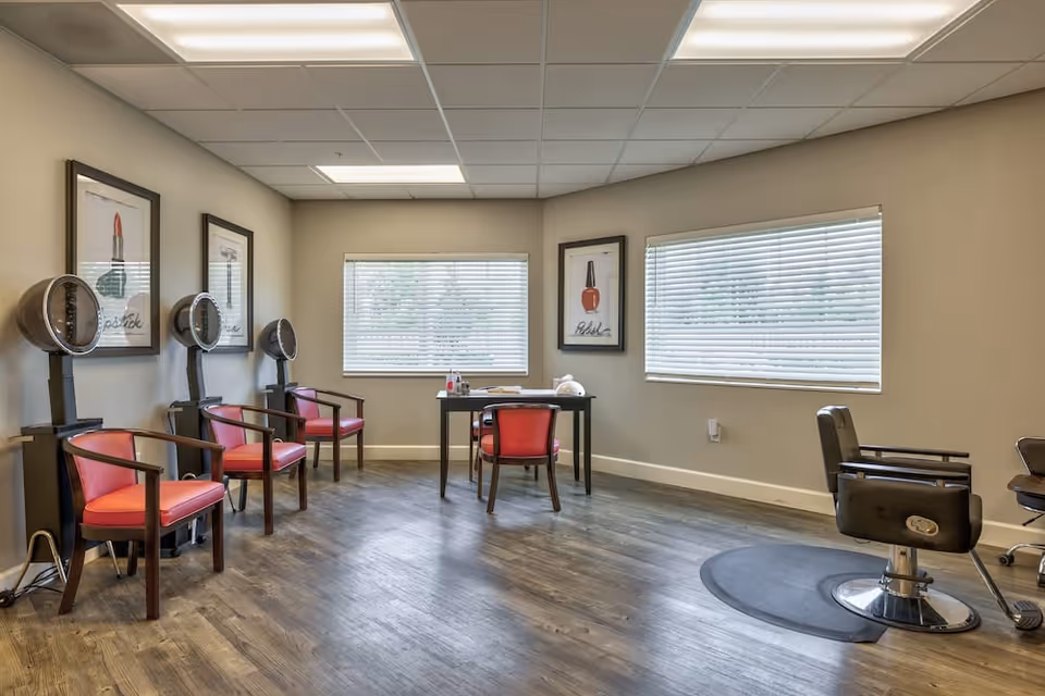 Interior of a salon area in a senior living facility with three red chairs under hair dryers on the left, a black table with a red chair in the center near two windows with blinds, and a black salon chair on the right on a protective mat. The walls are beige with framed pictures of lipstick and nail polish.