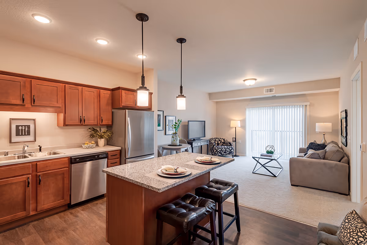 Open concept living space featuring a kitchen with wooden cabinets, stainless steel appliances, and a granite countertop island with two black cushioned stools. The living room area has a gray sofa, a patterned armchair, a glass coffee table, a TV on a stand, and large vertical blinds covering a sliding glass door.