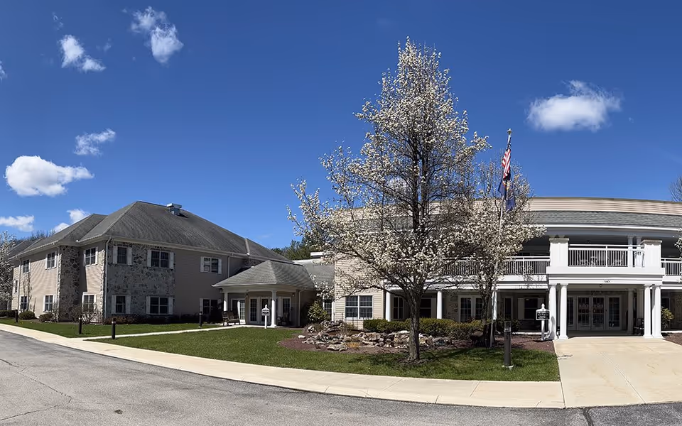 Exterior view of Westminster Woods senior living facility on a clear sunny day with a blue sky and a few clouds. The building has a beige and stone facade with a two-story section featuring white columns and a balcony. There is a blooming tree with white flowers in front of the entrance, along with a small landscaped area with rocks and shrubs. Two flagpoles with American and state flags are visible near the entrance.
