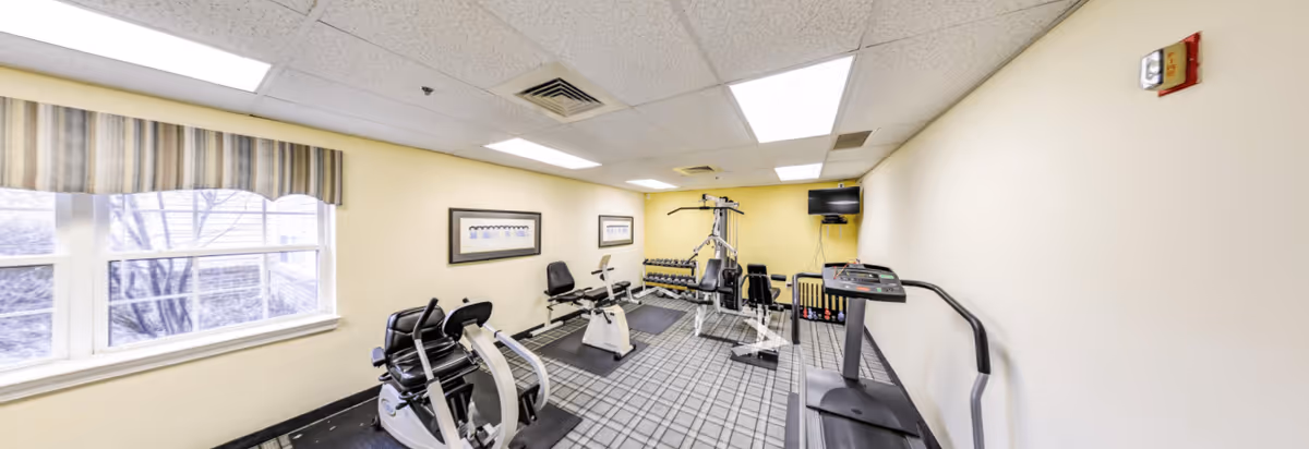 A small fitness room with exercise equipment including a treadmill, stationary bikes, a multi-function weight machine, and dumbbells on a rack. The room has a window with a striped valance, framed pictures on the wall, and a wall-mounted TV. The ceiling has fluorescent lighting and the walls are painted light yellow.