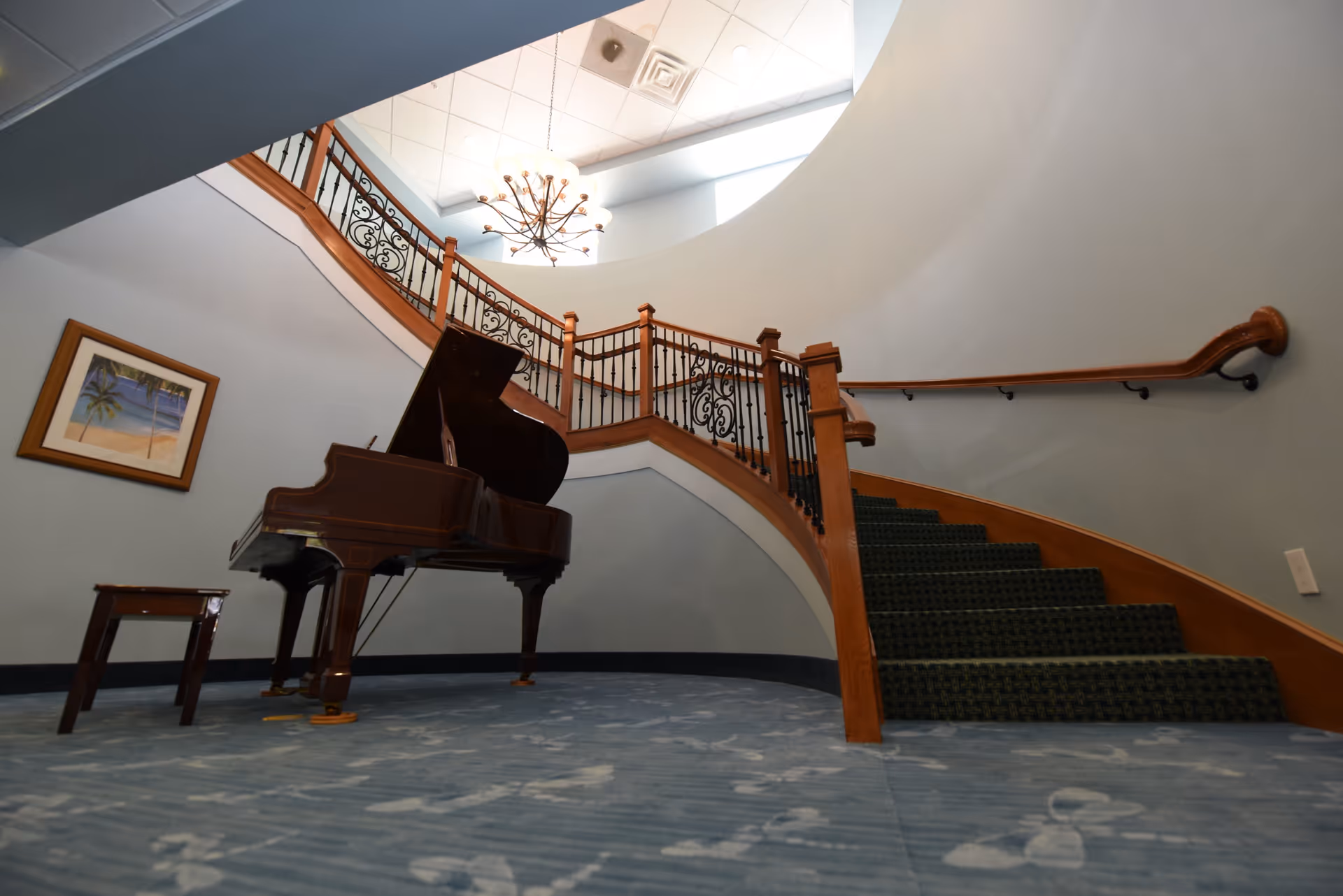Interior view of a curved staircase with wooden handrails and black wrought iron balusters. A grand piano with a matching bench is placed on the carpeted floor near the staircase. A framed painting of a beach scene hangs on the wall. A chandelier hangs from the ceiling above the staircase.