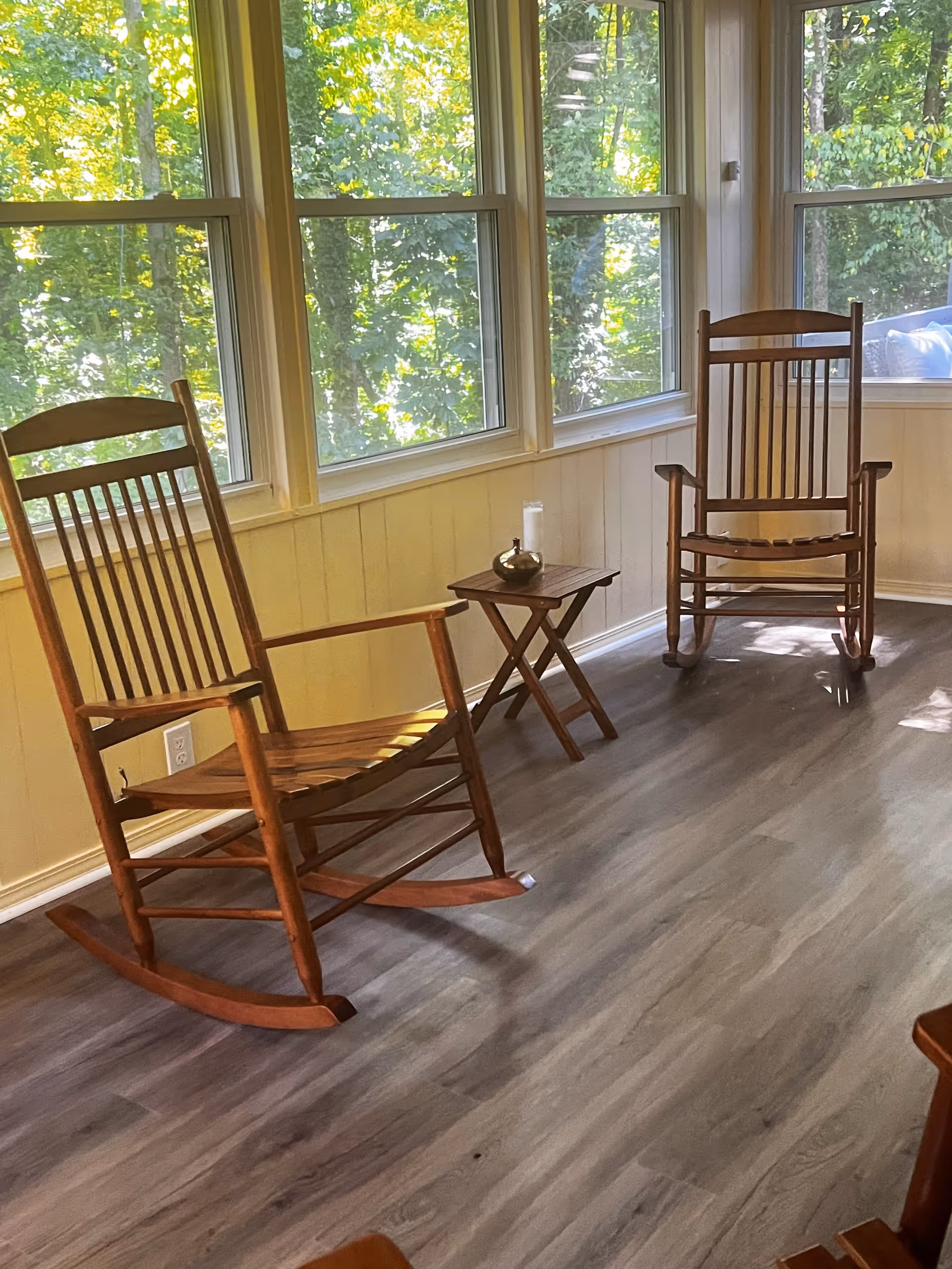 A bright sunroom with large windows showing green trees outside. Inside, there are two wooden rocking chairs and a small wooden side table with a candle and a decorative item on it. The floor is a light gray wood laminate.