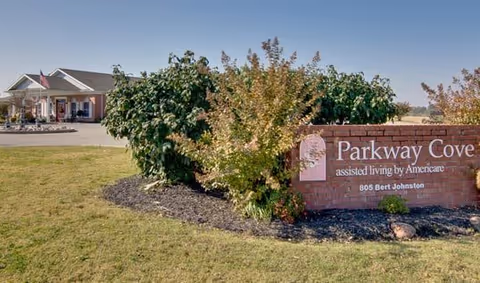 Exterior view of Parkway Cove assisted living facility with a brick sign displaying the facility name and address, surrounded by bushes and a lawn, with the building and entrance visible in the background under a clear sky.