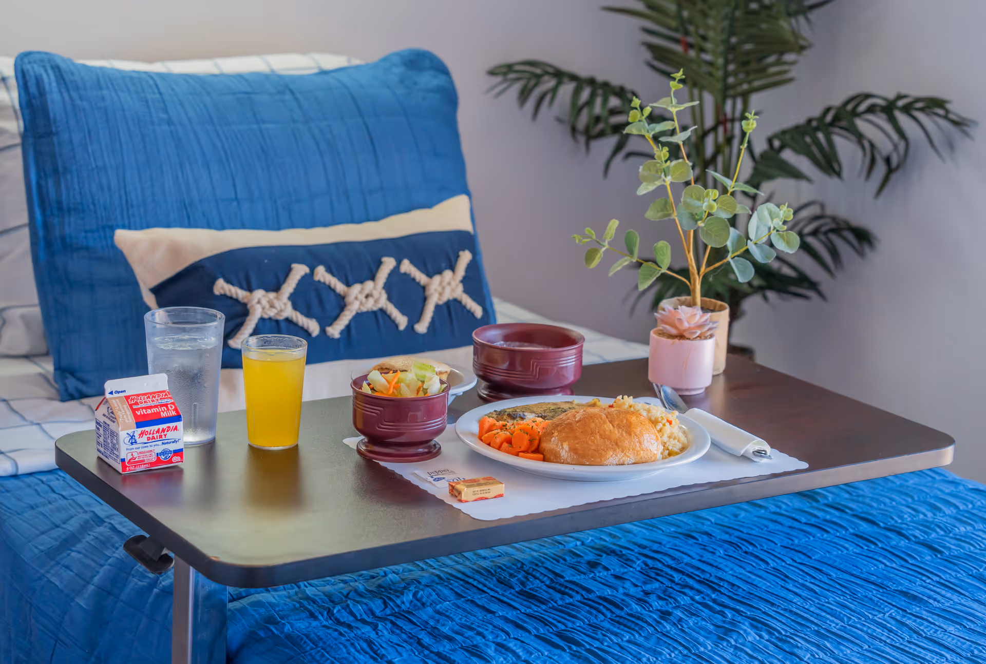 A plated meal with salad, vegetables, a roll and drinks served on a tray over a bed with blue bedding.
