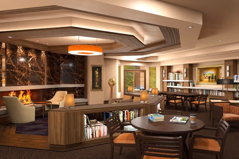 A cozy and well-lit common area with a modern fireplace, comfortable armchairs, round tables with chairs, bookshelves filled with books, a globe, and warm lighting fixtures. The space has a wooden ceiling design and large windows letting in natural light.