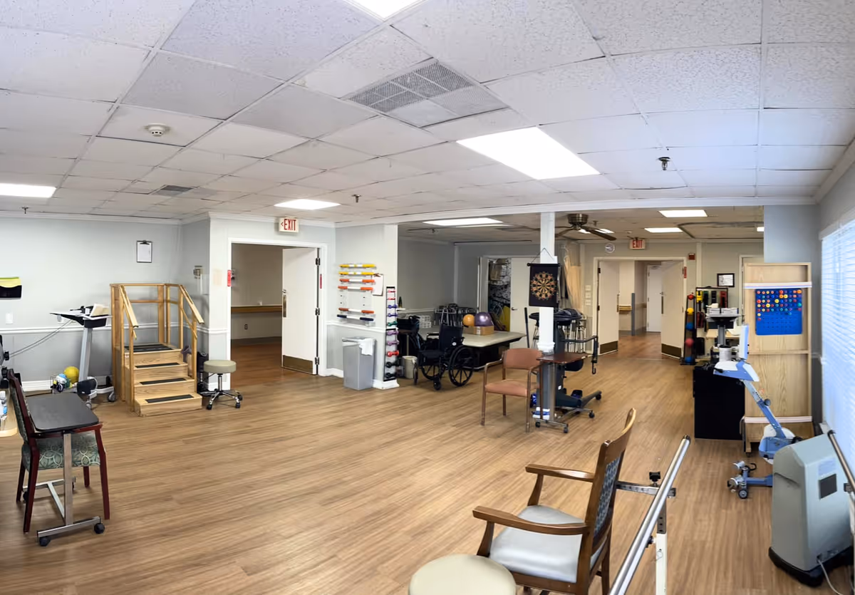 A spacious rehabilitation room with wooden flooring and white walls. The room contains various physical therapy equipment including parallel bars, a small staircase for stair training, exercise balls, a wheelchair, and a dartboard on the wall. There are chairs and stools scattered around, and the ceiling has fluorescent lighting panels. Doors and hallways lead to other parts of the facility.