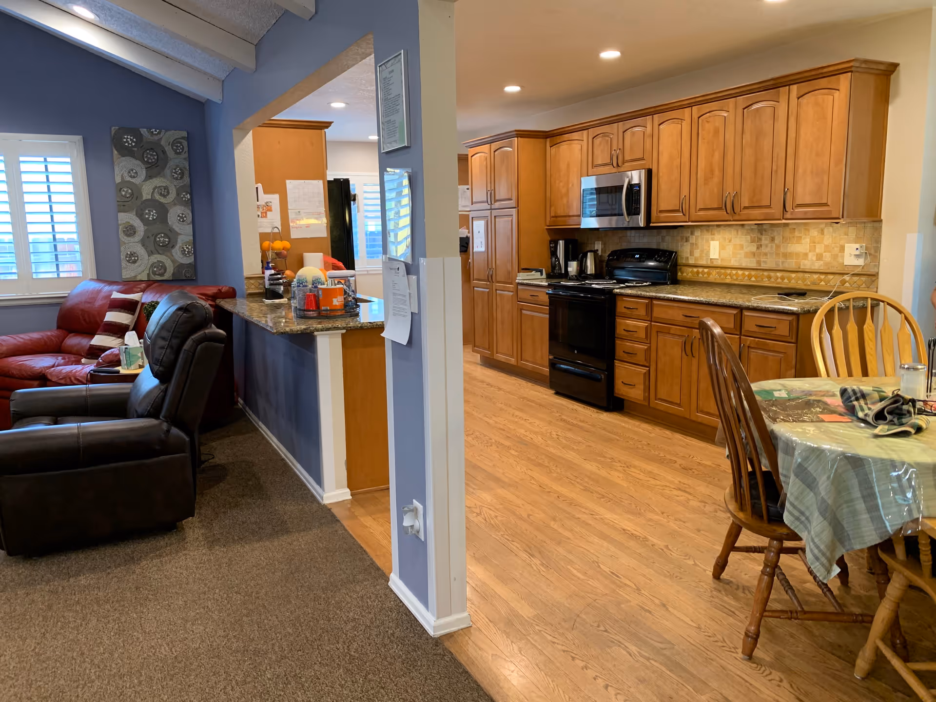 Interior view of a senior living facility showing a cozy living room area with a red leather couch and a black recliner chair on carpeted flooring. Adjacent to the living room is a kitchen with wooden cabinets, a black stove, microwave, and granite countertops. A dining table with a checkered tablecloth and wooden chairs is partially visible on the right side. The walls are painted in blue and beige tones, and there is a decorative wall hanging above the couch.