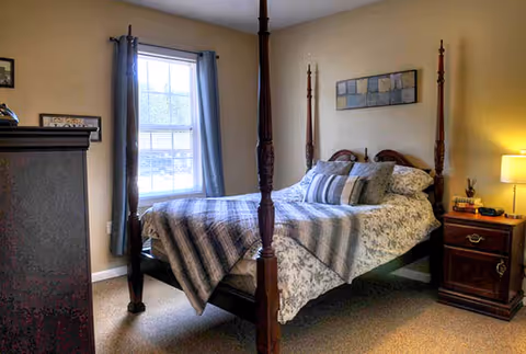 A cozy bedroom with a four-poster bed covered in patterned bedding and multiple pillows. There is a nightstand with a lamp and some items on it to the right of the bed. A window with blue curtains is on the left wall, and a dresser is partially visible in the foreground on the left side. The walls are painted a light beige color.