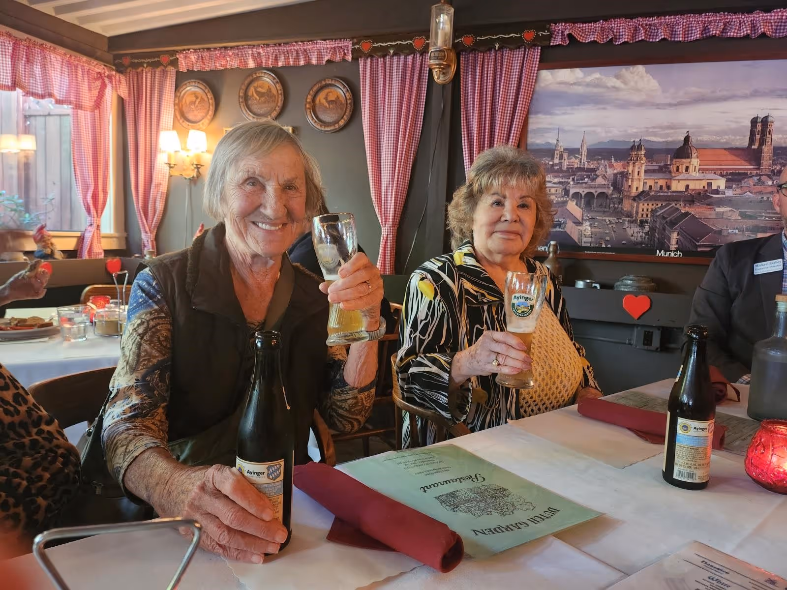Two smiling older adults sit at a decorated dining table holding beer glasses in a cozy dining room.