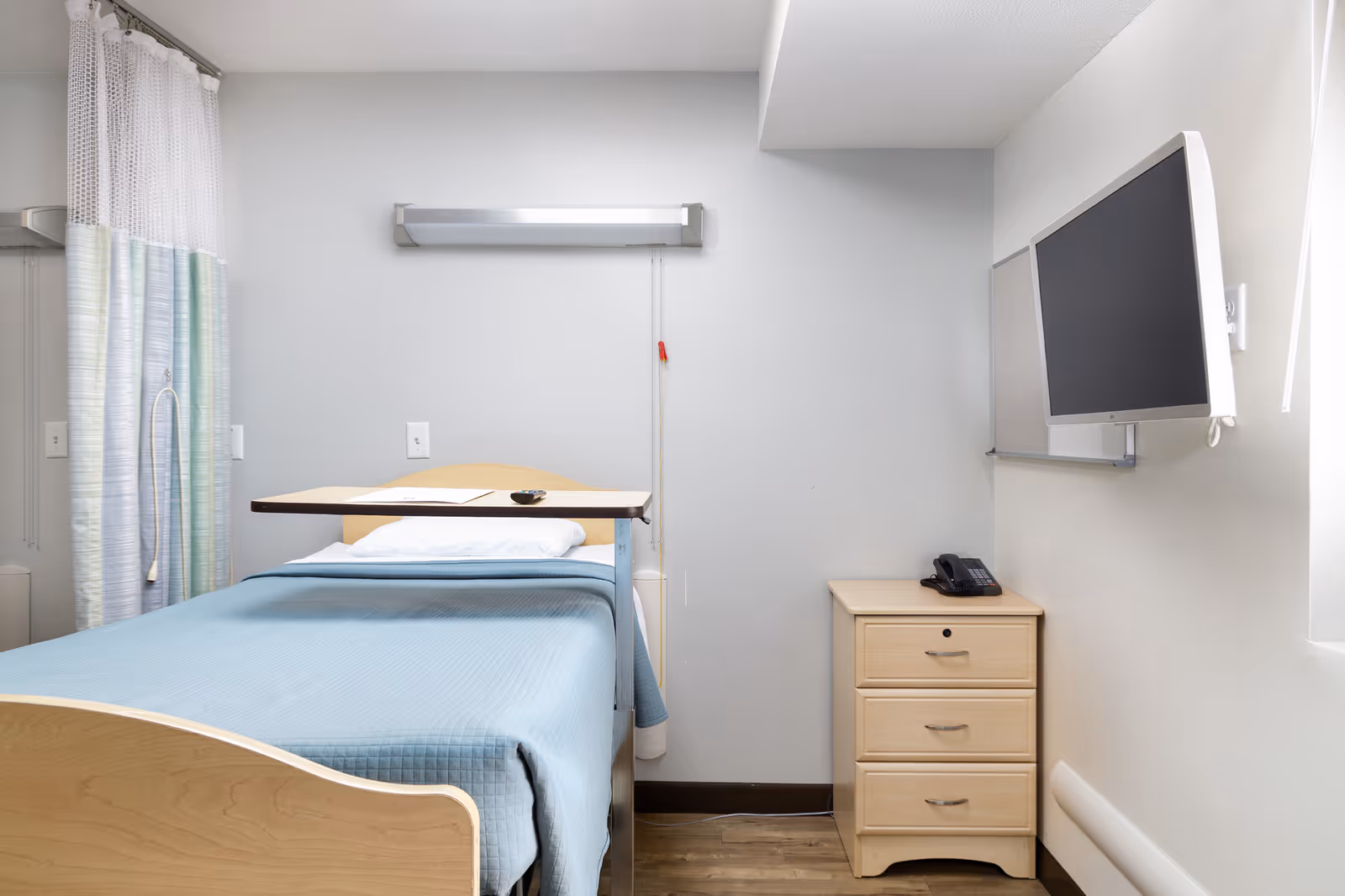 A simple hospital-style room with a single bed covered in a blue blanket and a white pillow. There is an overbed table positioned over the bed. To the right of the bed is a small wooden nightstand with three drawers and a black telephone on top. A flat-screen TV is mounted on the wall above the nightstand. Light blue and white curtains are partially visible on the left side of the image.