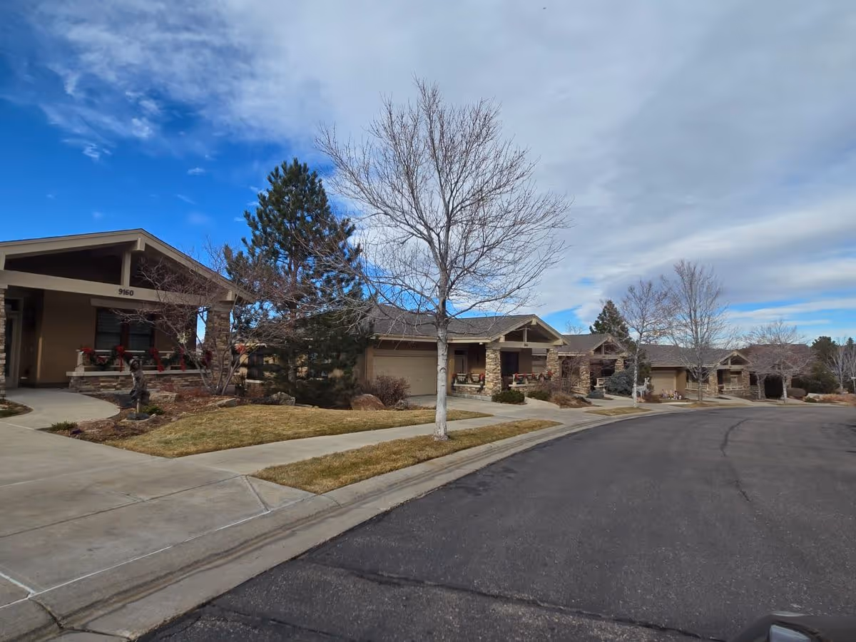 Row of single-story residence units with porches along a curved street, leafless trees, and a partly cloudy sky.