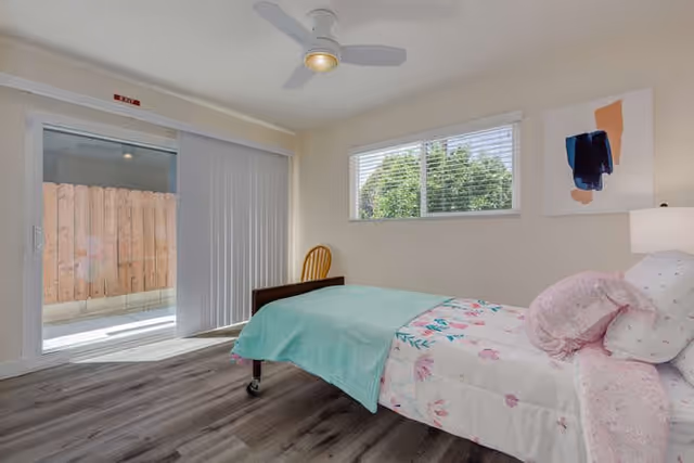Simple bedroom with a single bed covered in floral bedding, a ceiling fan, a sliding glass door, a window, and wood-look flooring.