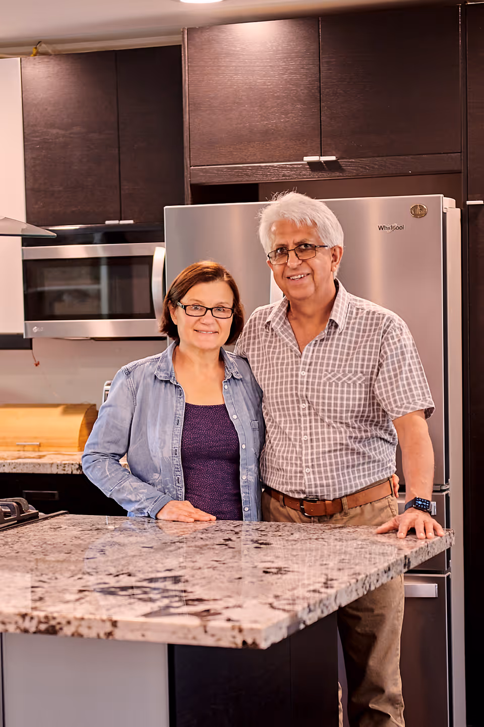 An older couple standing together in a modern kitchen with dark cabinets, a stainless steel refrigerator, and a granite countertop island. Both are smiling and wearing glasses.
