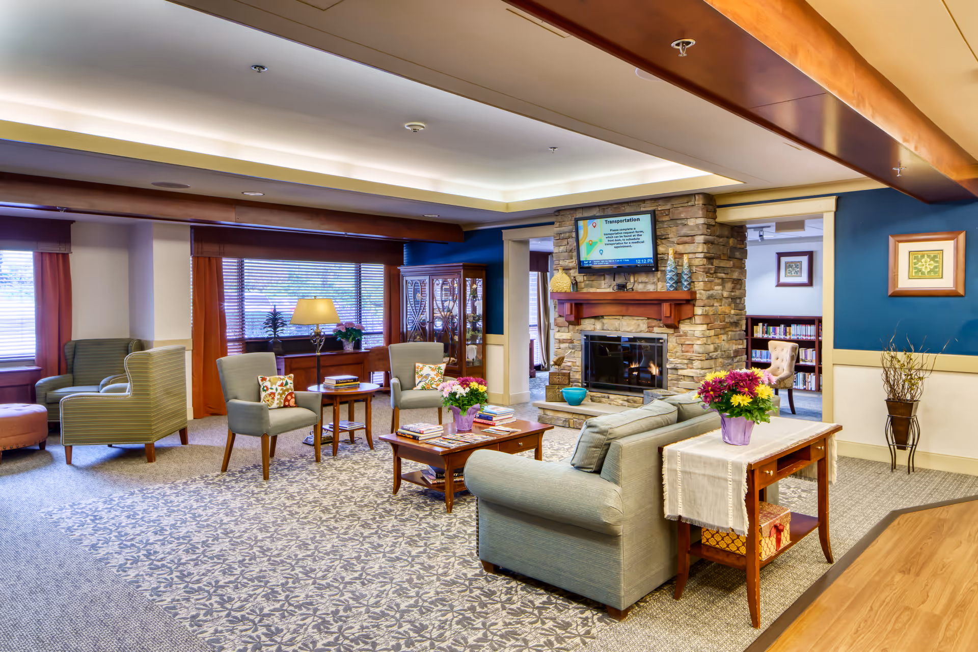 Spacious, well-lit common lounge with sofas and armchairs arranged around coffee tables facing a stone fireplace with a wall-mounted TV.