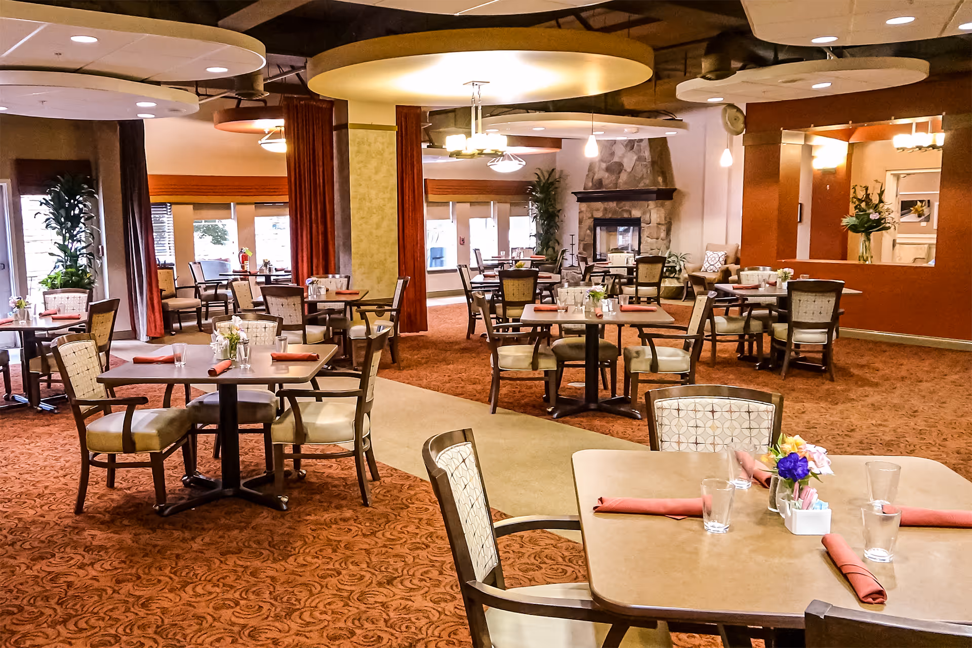 A spacious dining room in a retirement facility with multiple tables and chairs arranged neatly. Each table has a small flower arrangement and folded red napkins. The room features warm lighting, carpeted floors, large windows with curtains, and a stone fireplace in the background.