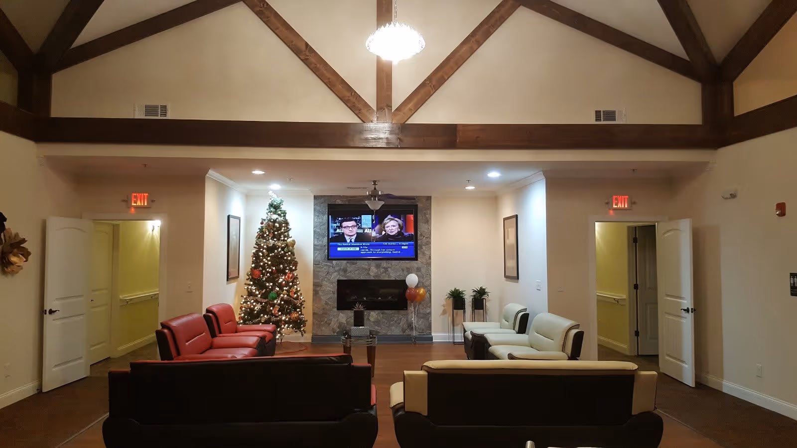 A spacious senior living common area with high vaulted ceilings and wooden beams. The room features a decorated Christmas tree, a wall-mounted flat-screen TV above a fireplace, and several sofas arranged for seating. There are two open doorways on either side of the room, and the space is warmly lit with a chandelier hanging from the ceiling.