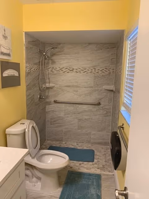 Small accessible bathroom with a toilet, open tiled shower with a grab bar and handheld showerhead, yellow walls, and blue bath mats.