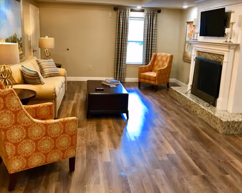 Sunlit living room with a sofa, two patterned armchairs, a dark coffee table, and a fireplace with a wall-mounted TV.