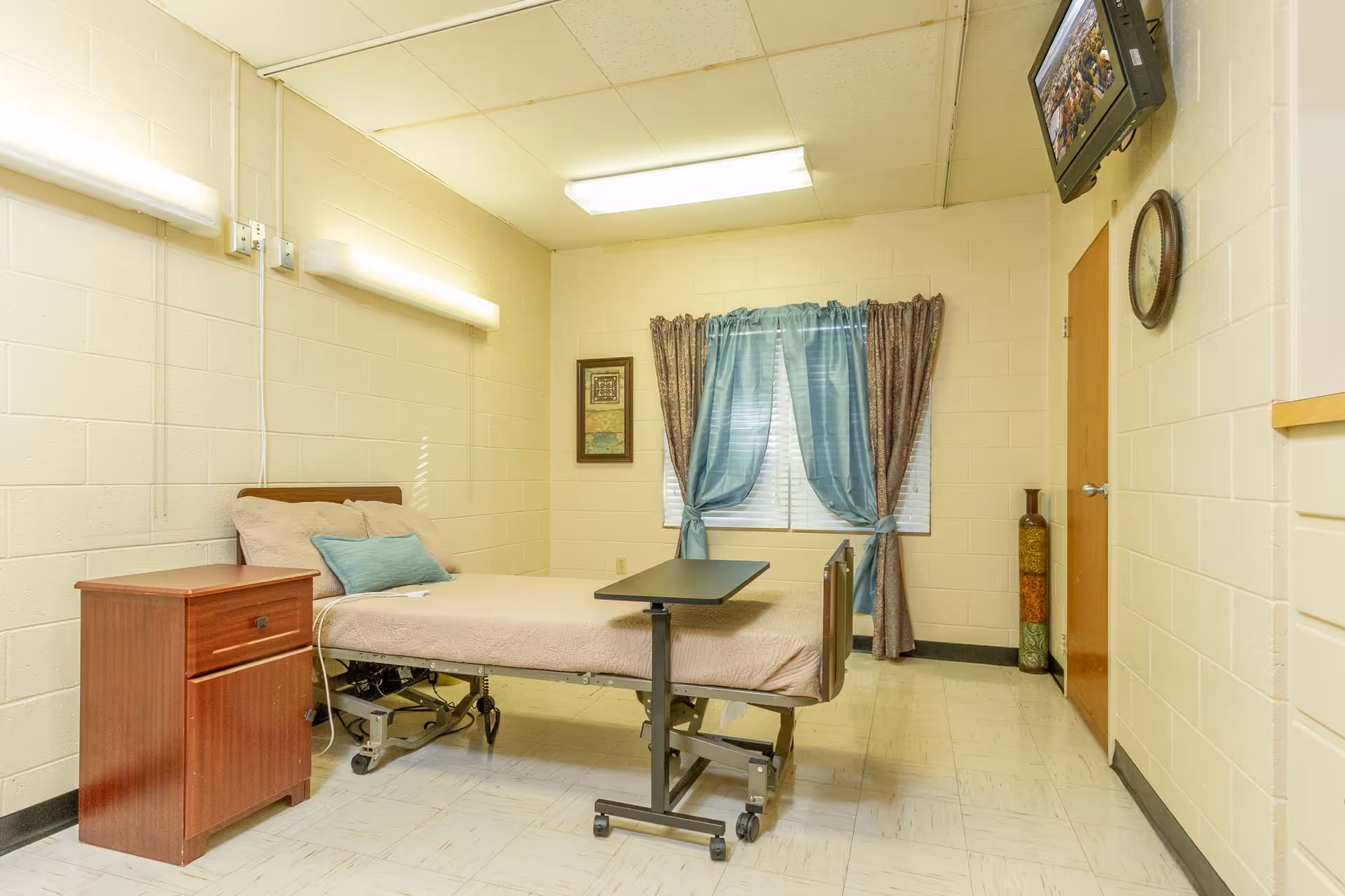 A simple nursing facility bedroom with a hospital bed, a wooden nightstand, a small overbed table, a window with blue and brown curtains, a wall-mounted TV, a clock, and a decorative vase in the corner.