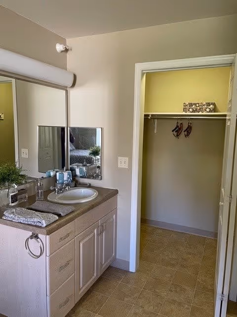 Bathroom vanity area with a sink, countertop, and mirror. The countertop has a soap dispenser, a small plant, and folded towels. To the right, there is an open doorway leading to a closet with a shelf and hanging hooks. The floor is tiled and the walls are painted light beige.