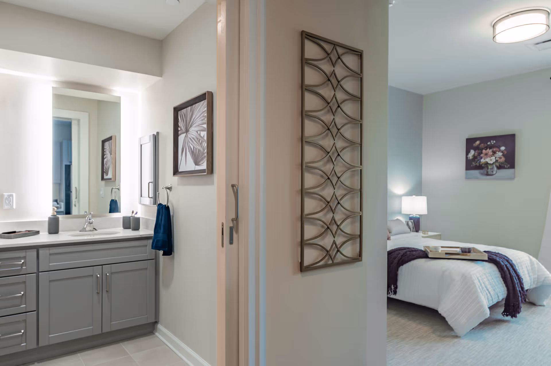 View of a bathroom vanity with a large mirror and gray cabinets on the left, and a bedroom with a bed, nightstand, lamp, and floral wall art on the right, separated by a partial wall with decorative metal artwork.