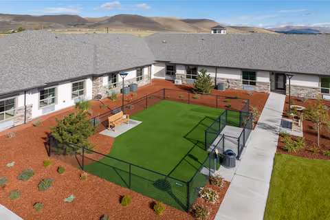 Aerial view of a fenced outdoor courtyard area at Kiley Ranch Senior Living, featuring a green artificial turf lawn, a wooden bench on a concrete pad, surrounding mulch beds with small plants and shrubs, and a paved walkway leading to the building entrances. The building has a gray shingled roof and light-colored walls with stone accents. Hills and a blue sky with clouds are visible in the background.