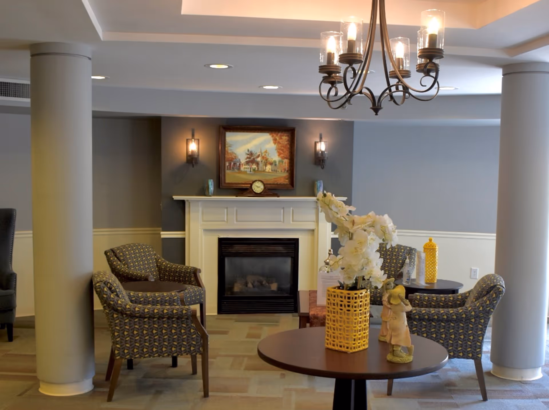A cozy sitting area with patterned armchairs arranged around a fireplace. Above the fireplace is a framed painting and a small clock. A round table in the foreground holds a yellow vase with white flowers and a small figurine. The room has soft lighting with wall sconces and a chandelier hanging from the ceiling.