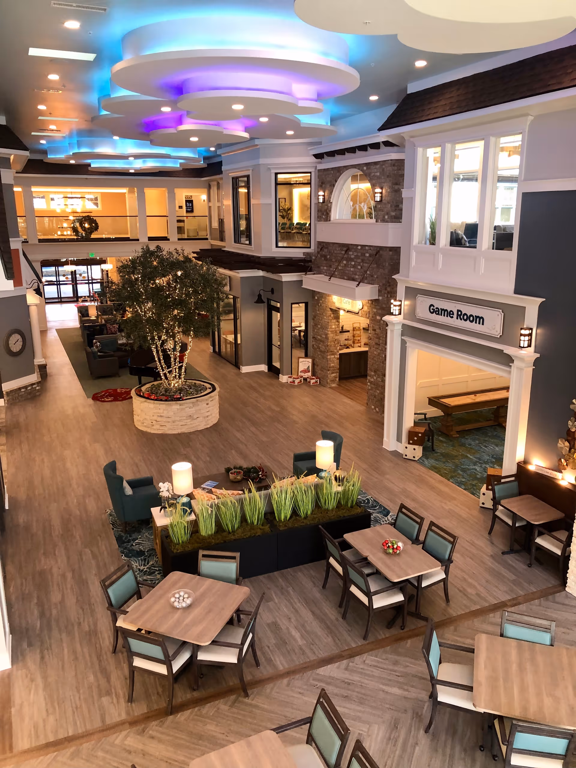 Interior view of a senior living facility common area with modern decor, featuring tables and chairs arranged for socializing, a central planter with green plants, comfortable seating areas, and a game room entrance. The ceiling has circular light fixtures with blue and purple lighting accents.
