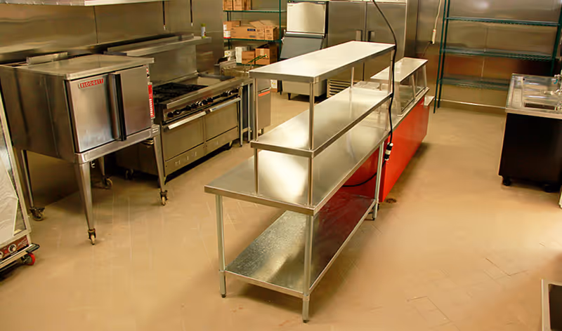 Commercial kitchen area with stainless steel appliances including an oven, stove, refrigerator, and metal shelving units on a tiled floor.