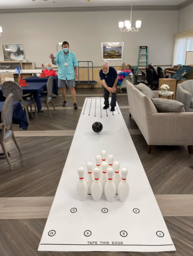 An elderly man is bowling on a miniature indoor bowling lane set up in a living room area. A staff member wearing a mask and teal shirt stands nearby, and two other elderly individuals are seated in the background. The room has chairs, tables, and a couch, with framed pictures on the walls and a chandelier overhead.