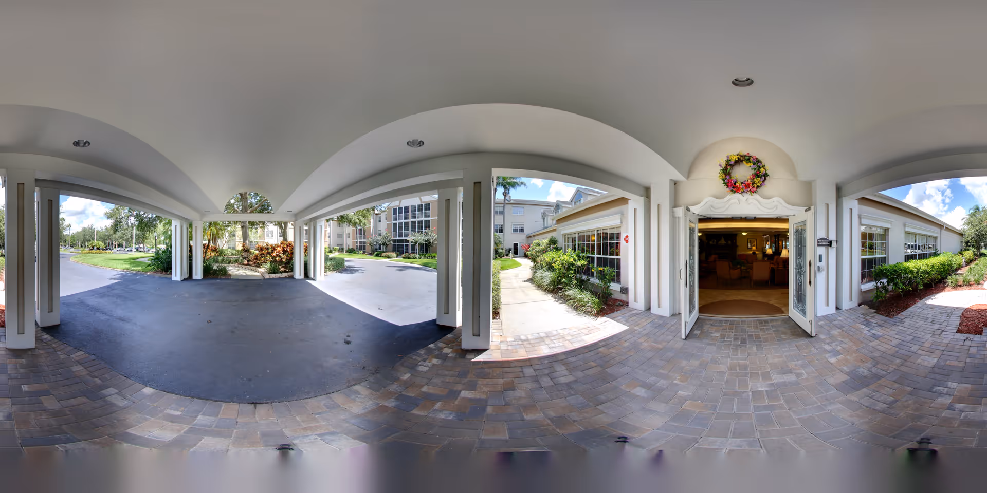 Covered porte-cochere and main entrance of a senior living building with columns, tiled driveway, landscaping, and open double doors into the lobby.