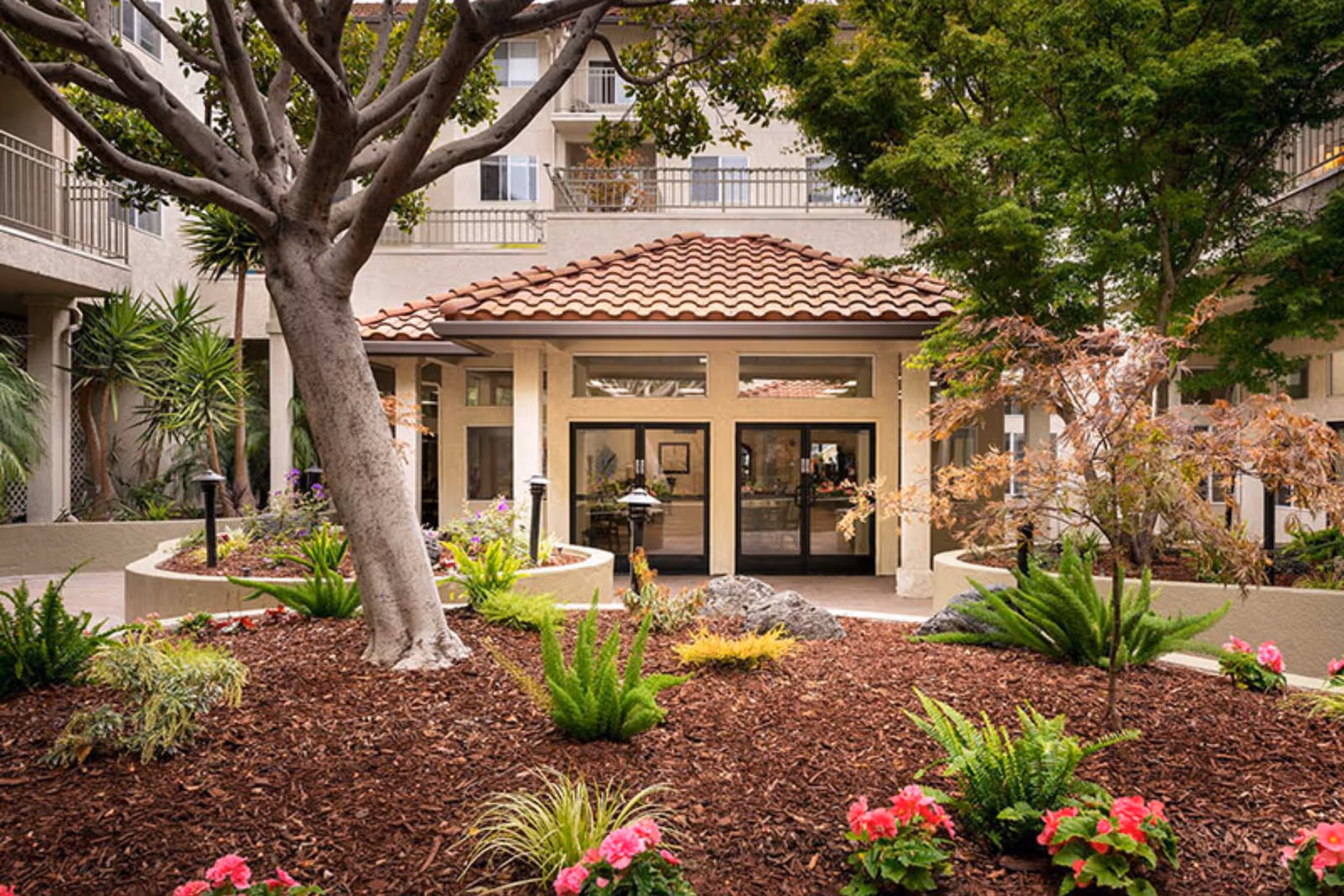 Landscaped courtyard with trees and flowers leading to the tile-roofed entrance of a senior living building.