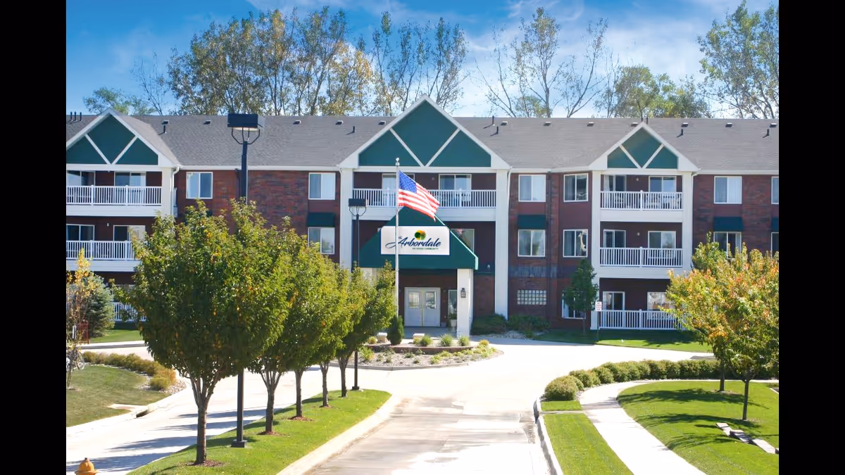 Front exterior of The Arbordale three-story brick senior living building with balconies, an American flag over the main entrance, a circular driveway and landscaped trees.