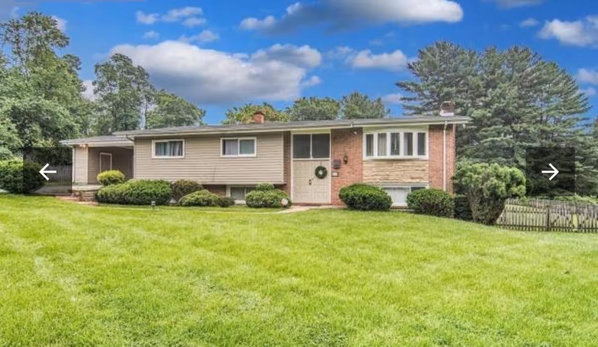 Single-story residential building with beige siding and brick accents, surrounded by green bushes and a well-maintained lawn under a partly cloudy blue sky.