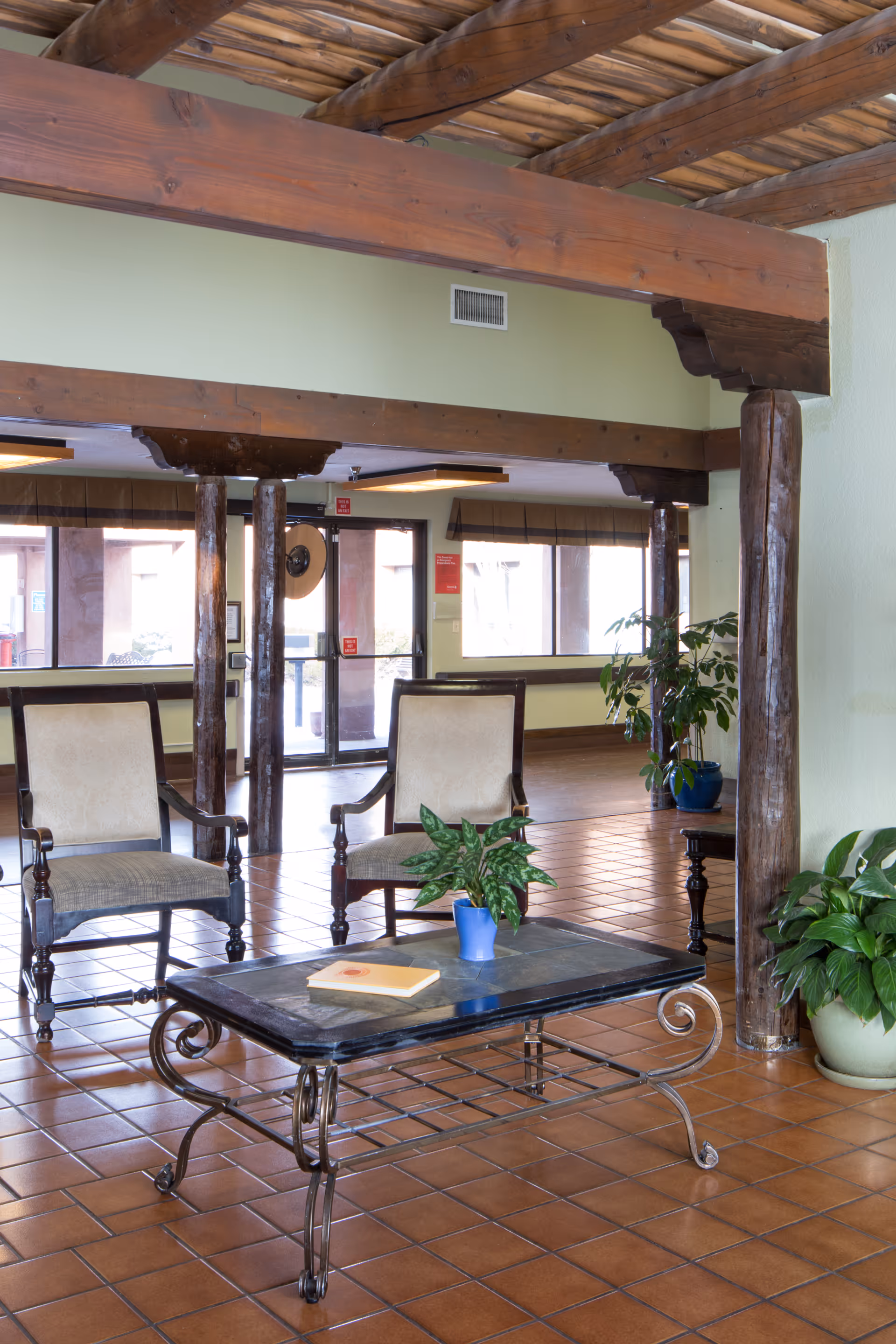 A cozy seating area in a senior living facility with two cushioned wooden chairs and a black metal coffee table with a potted plant and a book on it. The room features terracotta tile flooring, wooden beams on the ceiling, and large windows letting in natural light. There are additional potted plants near the wooden columns and walls painted in a light green shade.