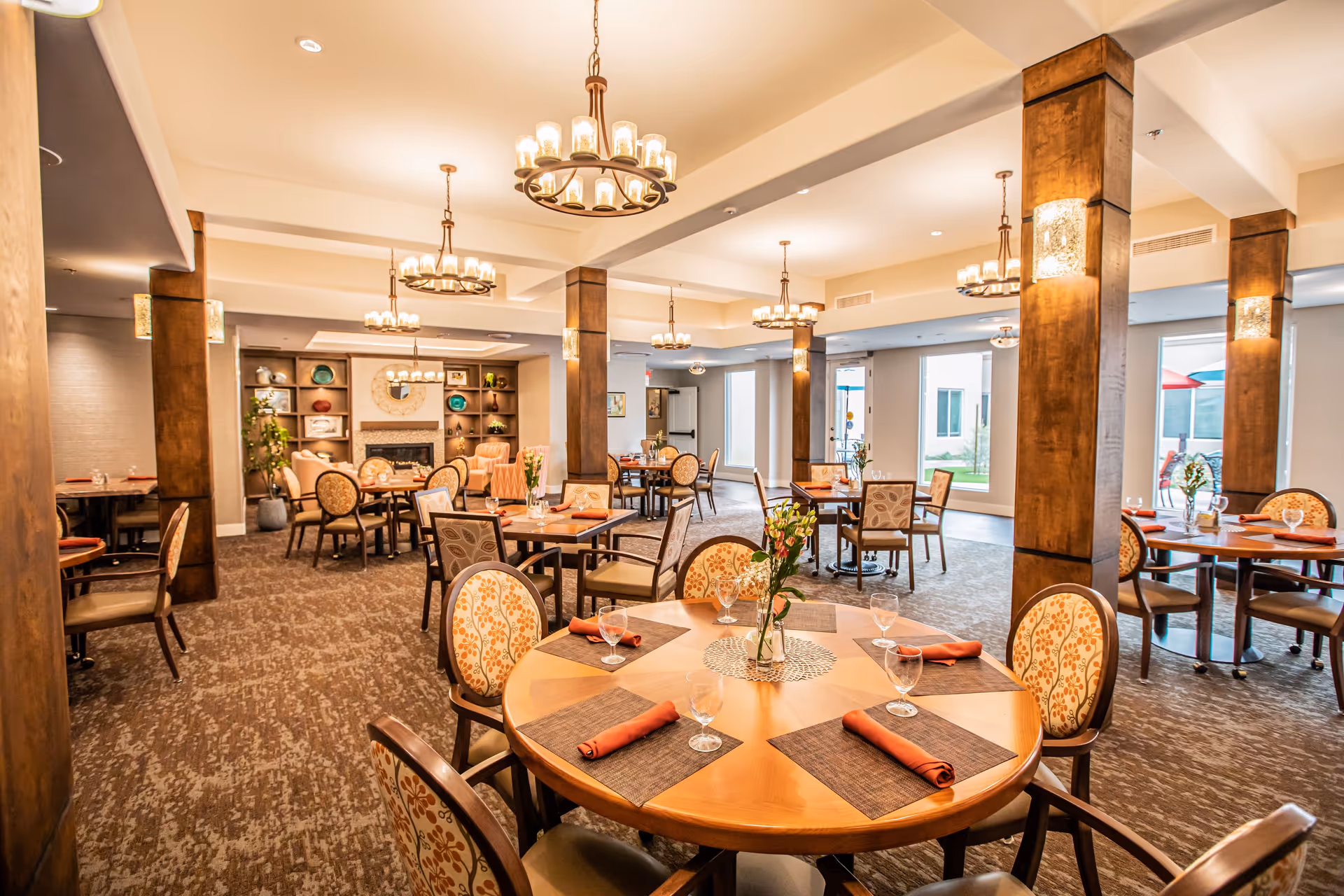 A spacious and well-lit dining room with multiple round and square wooden tables set with placemats, rolled orange napkins, and water glasses. The room features patterned upholstered chairs, wooden columns, chandeliers with multiple lights, and large windows letting in natural light. In the background, there is a cozy seating area with armchairs and a fireplace surrounded by shelves with decorative items.