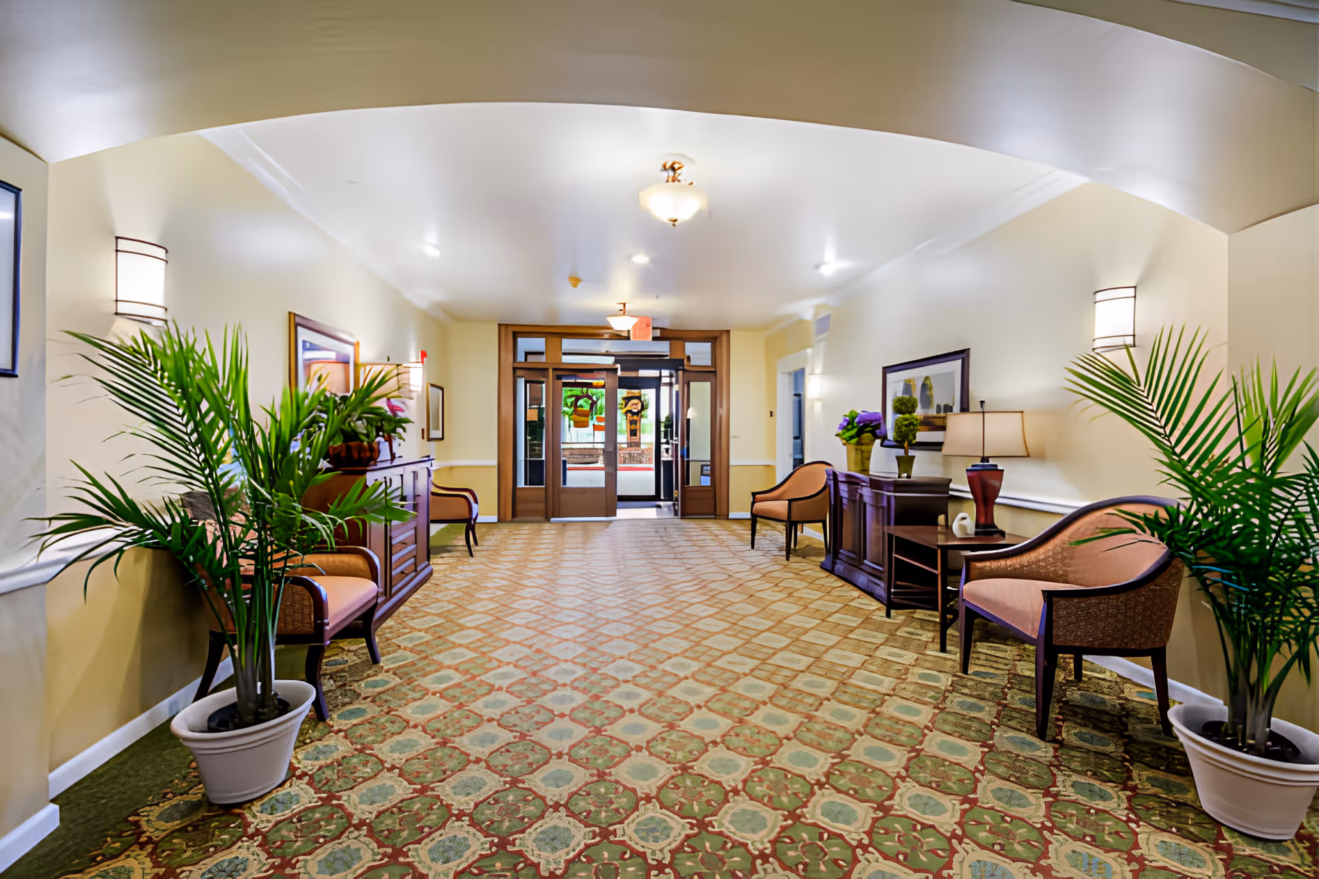 A carpeted lobby hallway with seating, potted plants, side tables and double glass entrance doors at the far end.