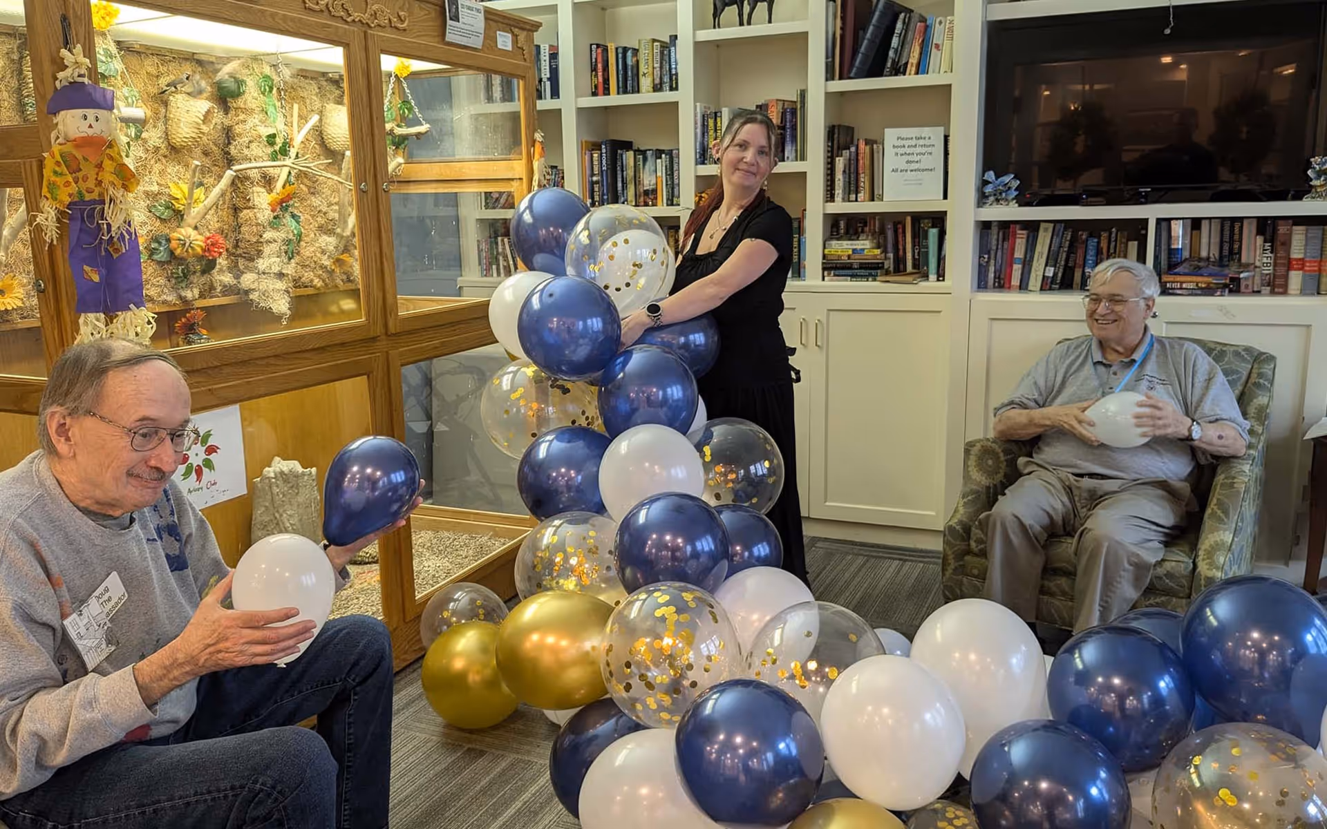 Two elderly men and a woman in a room decorated with bookshelves and a display cabinet. The woman is holding a large bunch of blue, white, gold, and transparent balloons with gold confetti inside. One man is sitting on the floor holding balloons, and the other man is sitting in a chair also holding a balloon. The room has a cozy, home-like atmosphere with a carpeted floor and a TV mounted on the wall.