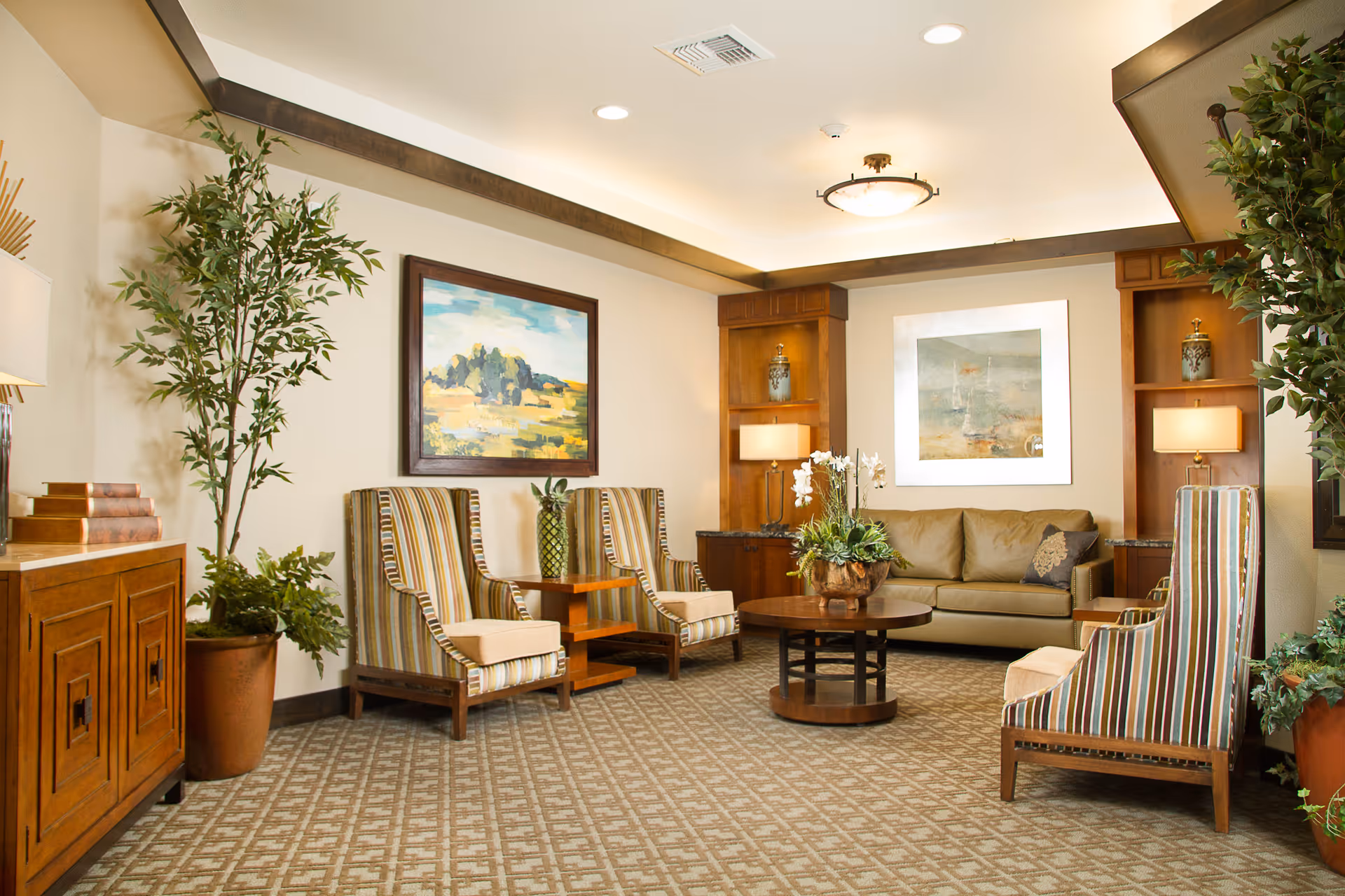 A cozy living room area with a beige sofa, four striped armchairs, a round wooden coffee table with a floral arrangement, two large potted plants, wooden cabinets with lamps, and framed landscape paintings on the walls.