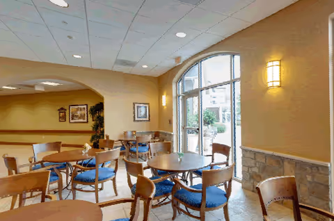 A bright dining area with several round wooden tables and chairs with blue cushions. Large arched windows allow natural light to fill the room, and the walls are painted a warm beige color with stone accents along the lower portion. Wall-mounted lights provide additional illumination.