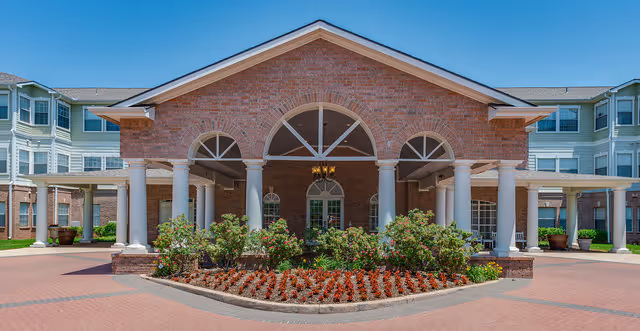 Front exterior view of a senior living facility named Town Village with a brick facade, white columns, arched windows, and a landscaped flower bed in front under a clear blue sky.
