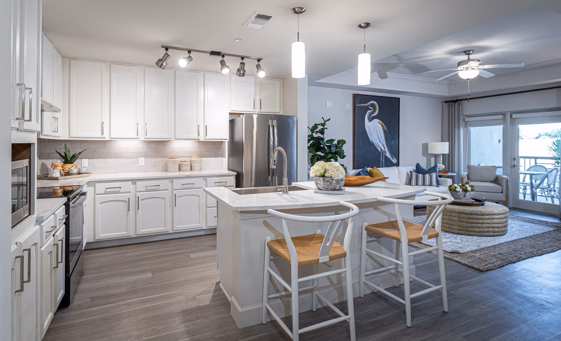 Modern open-concept kitchen and living room area with white cabinetry, stainless steel appliances, a kitchen island with two white chairs, pendant lights, and a living room featuring a white sofa, armchair, coffee table, large bird painting, ceiling fan, and glass doors leading to an outdoor patio.