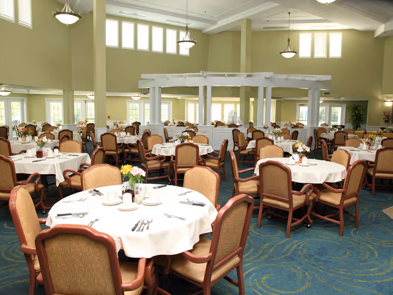 A spacious dining room with multiple round tables covered with white tablecloths, each set with plates, cups, utensils, and small flower arrangements. The room has high ceilings with hanging light fixtures and large windows allowing natural light to fill the space. The chairs are wooden with cushioned seats and backs.