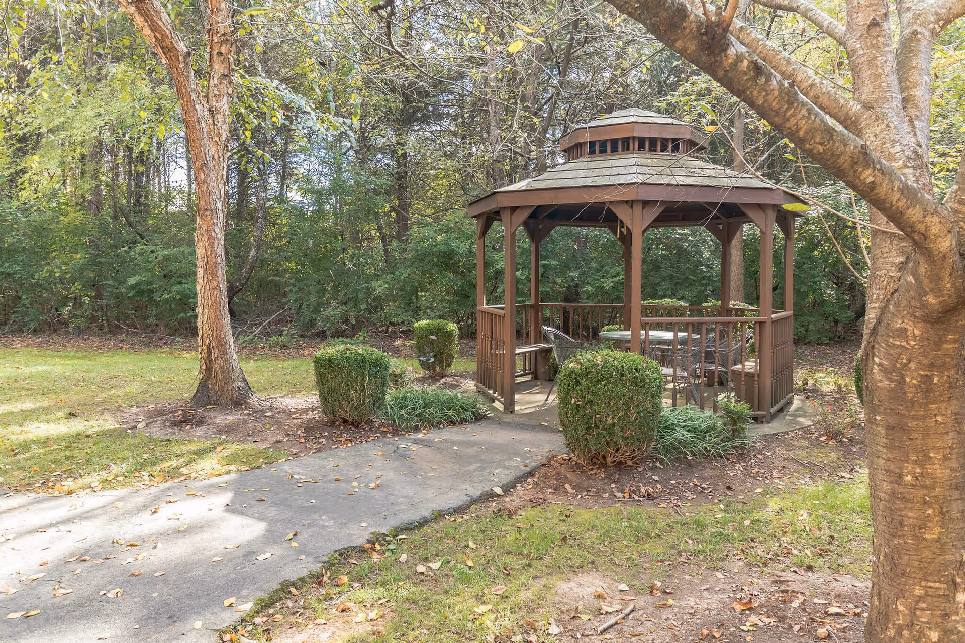 A wooden gazebo with a shingled roof situated in a garden area surrounded by trees and bushes. There is a paved pathway leading to the gazebo, which contains a table and chairs inside. The scene is outdoors with natural greenery and fallen leaves on the ground.