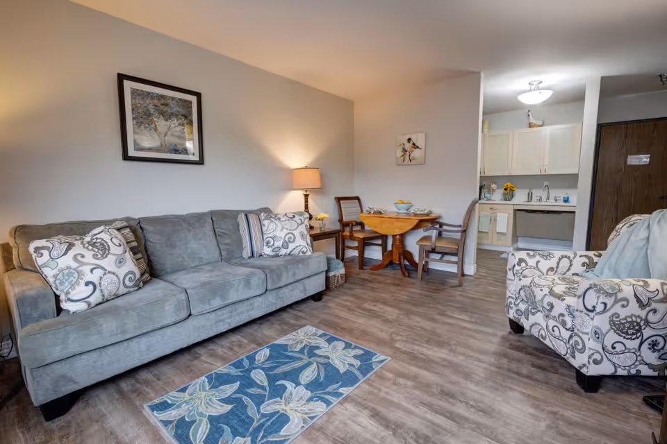 A cozy living area with a gray sofa adorned with patterned pillows, a floral armchair, a small wooden dining table with two chairs, and a kitchen area in the background. The room has wood flooring, a framed picture on the wall, a table lamp, and a blue floral rug on the floor.