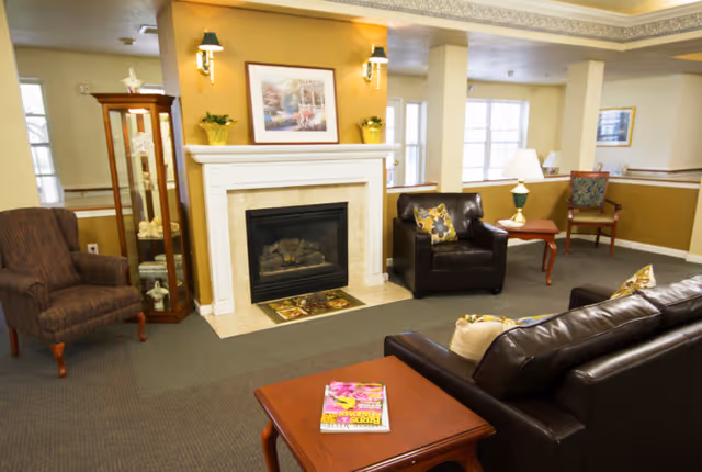 A cozy living room area with a fireplace centered on a yellow accent wall. The room features a dark leather sofa, a matching armchair with a floral pillow, a wooden side table with a lamp, and another armchair with a striped pattern. A glass display cabinet stands to the left of the fireplace, and framed artwork hangs above the mantle. The space is well-lit with natural light coming through windows in the background.