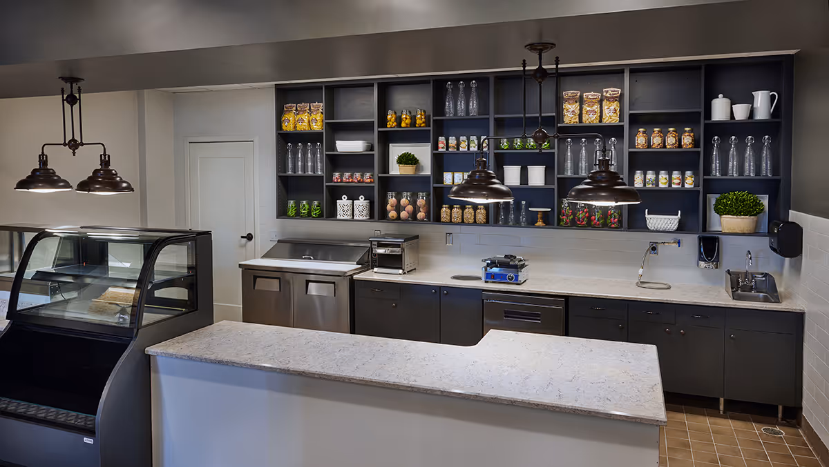 Modern kitchenette/cafe area with a marble-topped service counter, refrigerated display case, shelving with jars and glassware, and hanging pendant lights.