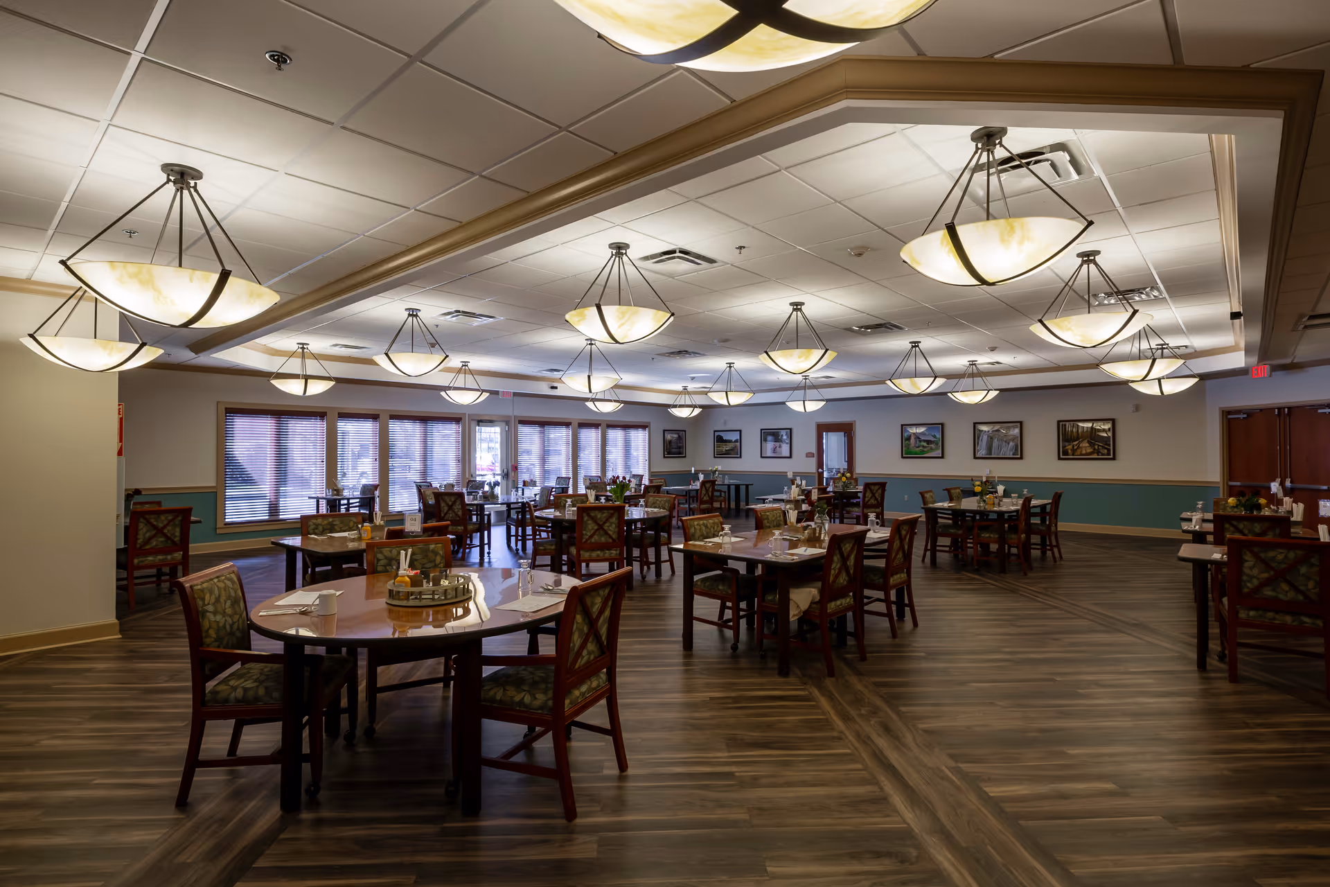 Spacious dining room with multiple round and rectangular tables, pendant ceiling lights, and large windows along one wall.