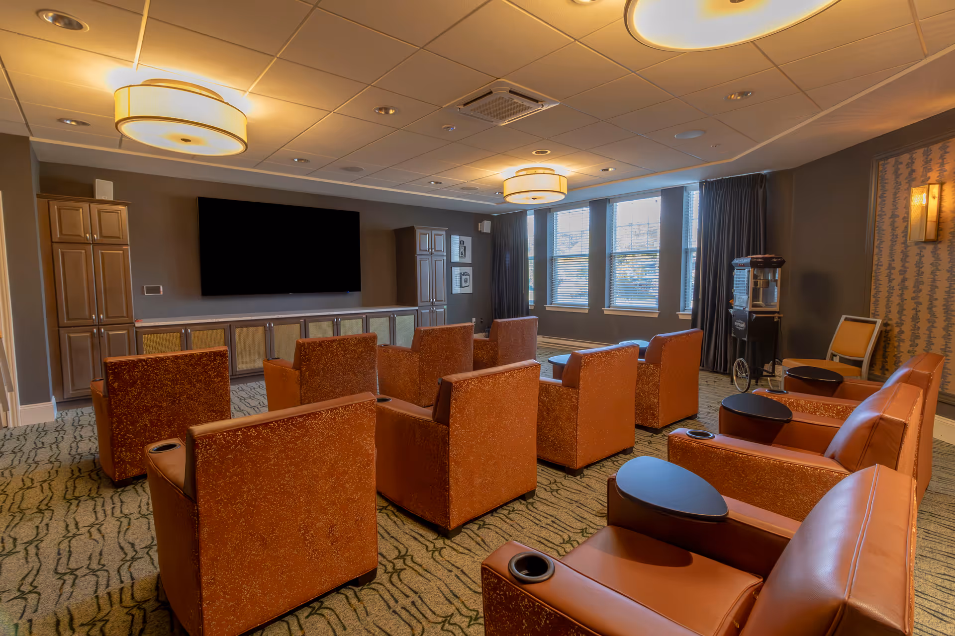 A cozy media room with multiple orange upholstered armchairs arranged in rows facing a large flat-screen TV mounted on the wall. The room features carpeted flooring, large windows with blinds, dark curtains, and soft ceiling lights. There is a popcorn machine in the corner and cabinets along the wall beneath the TV.